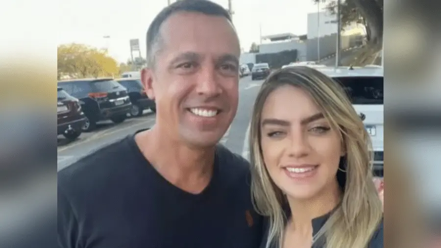 Imagem de um homem e uma mulher sorrindo em uma selfie ao ar livre, com carros de estacionamento ao fundo, sugerindo um momento alegre e descontraído.