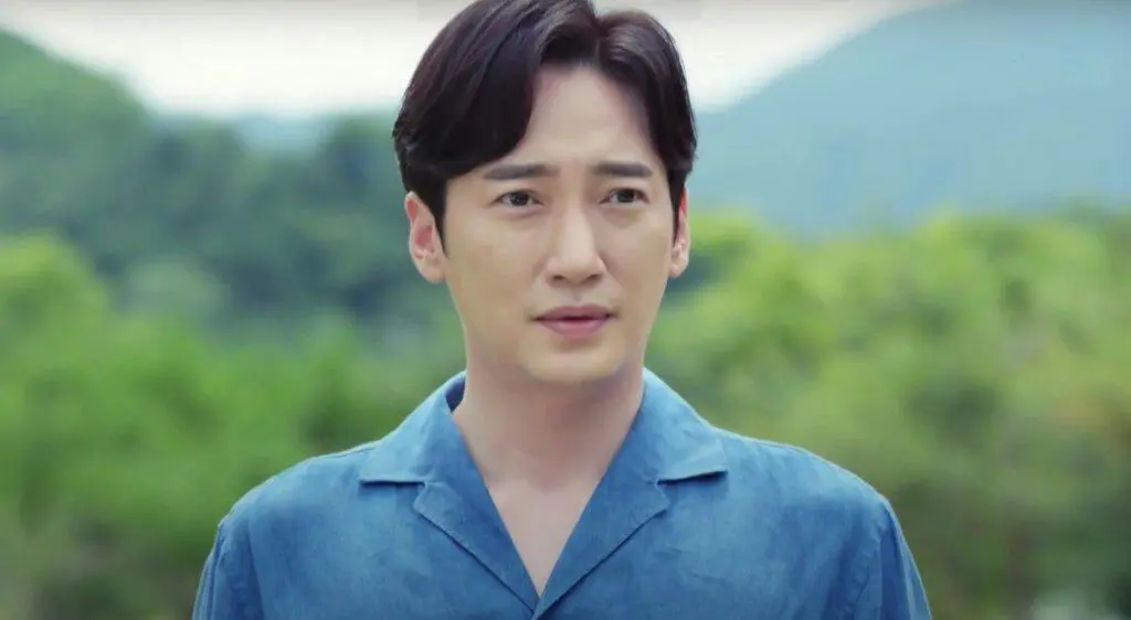 Lee Sang-Bo, um homem de cabelos escuros e expressão serena, veste uma camisa azul texturizada, posicionado em destaque contra um fundo natural e desfocado. A vegetação verde e as montanhas ao longe criam uma atmosfera tranquila e suave em torno do sujeito.