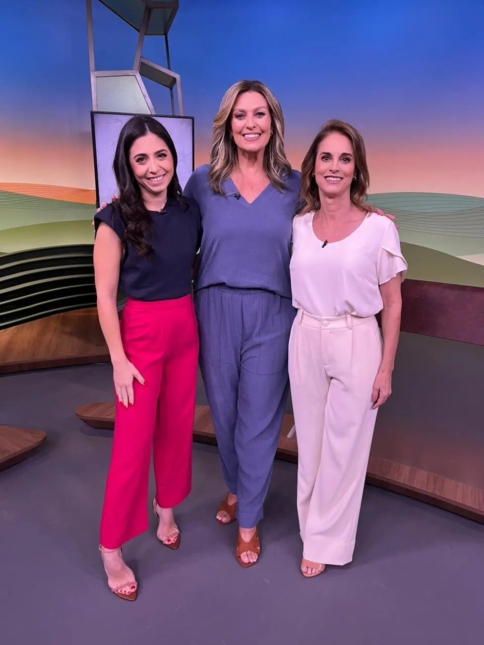 Três mulheres vestidas com roupas elegantes posam juntas em um estúdio de televisão, simbolizando uma equipe de jornalismo ou apresentadoras de TV.