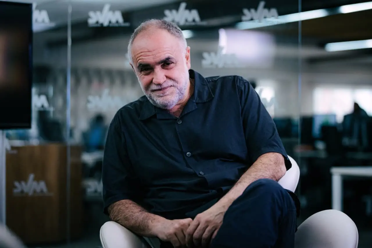 O cineasta Karim Aïnouz sorri levemente sentado em uma poltrona branca, vestindo uma camisa preta de botões em um ambiente de escritório moderno. Ao fundo, divisórias de vidro com logotipos e luzes suaves criam uma profundidade de campo que destaca sua expressão tranquila e experiente.