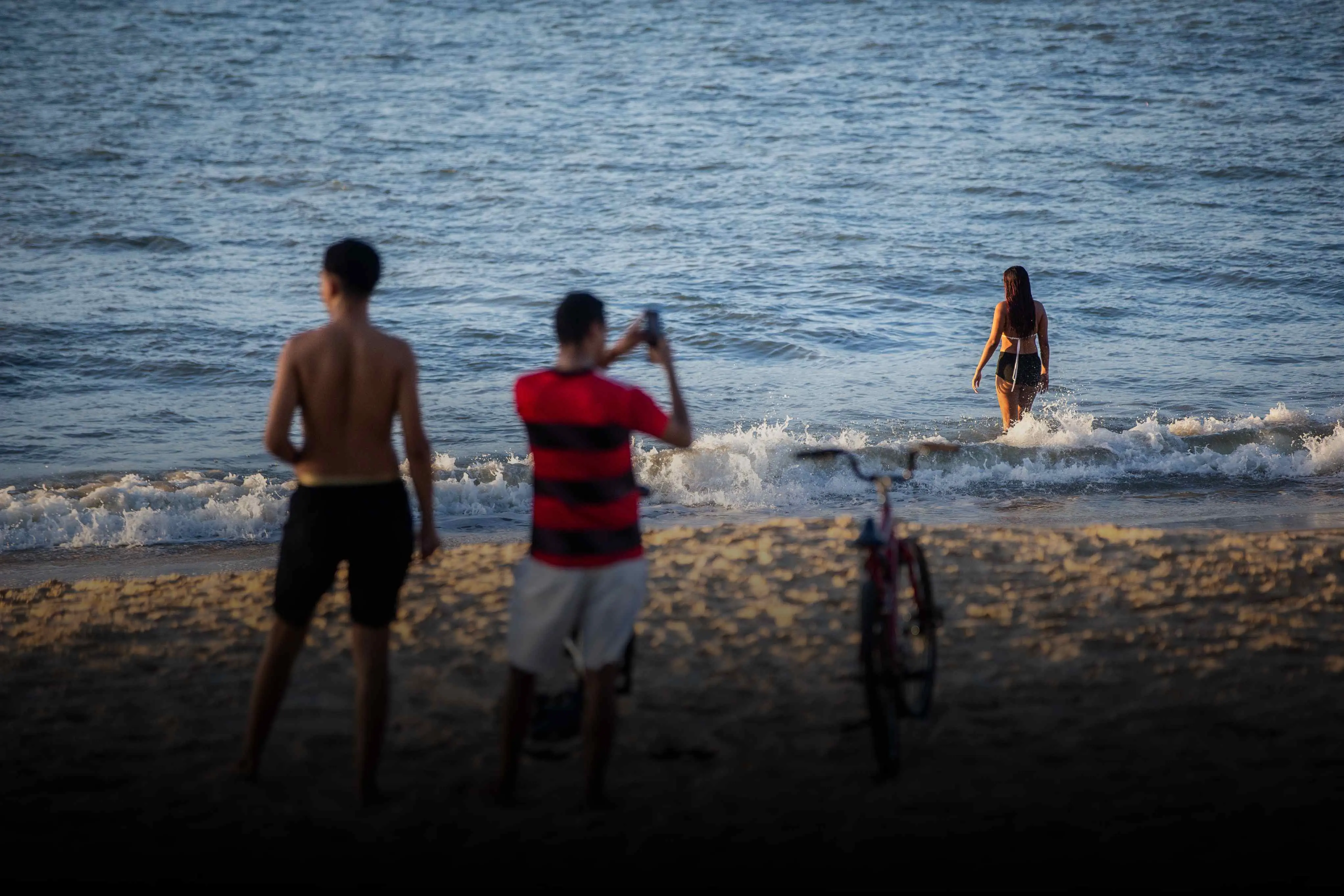 imagem na beira do mar mostra dois homens em primeiro plano, um deles segurando um celular. ao fundo, no mar, está uma mulher, também de costas.