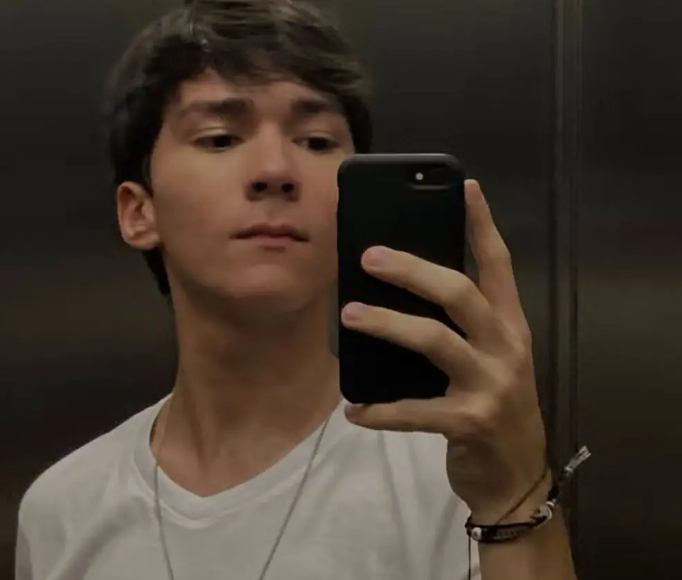 Pessoa tira foto no espelho de um elevador, usando camiseta branca e colar fino, segurando um celular preto; paredes metálicas ao fundo e pulseira no pulso.