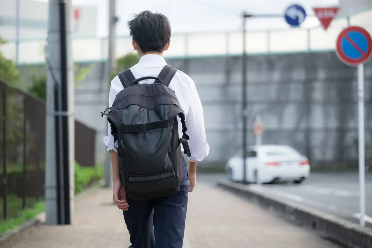 Imagem mostra estudante adolescente do gênero masculino de costas, andando por rua deserta, usando uniforme de blusa branca e mochila preta.