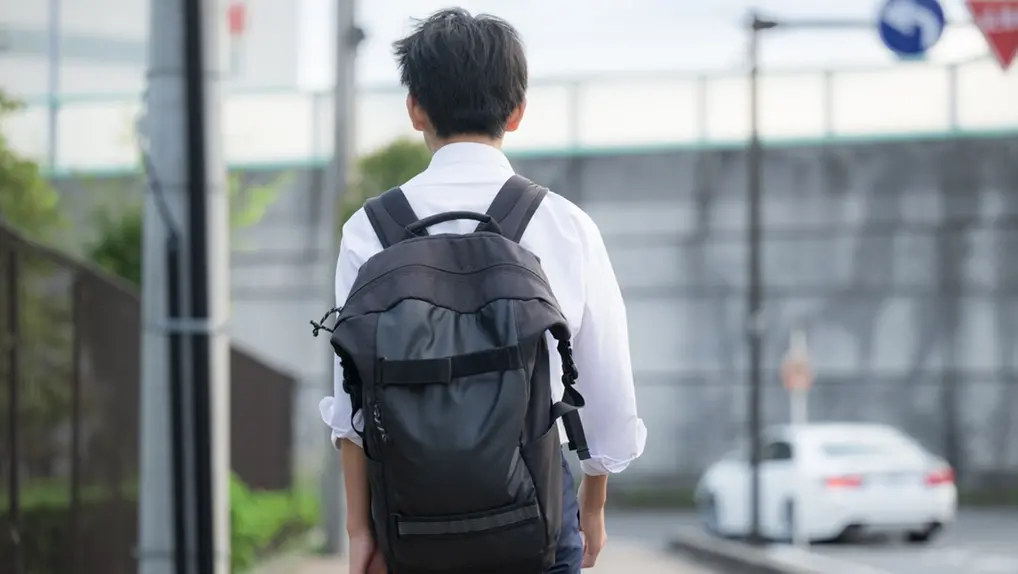 Imagem mostra estudante adolescente do gênero masculino de costas, andando por rua deserta, usando uniforme de blusa branca e mochila preta.