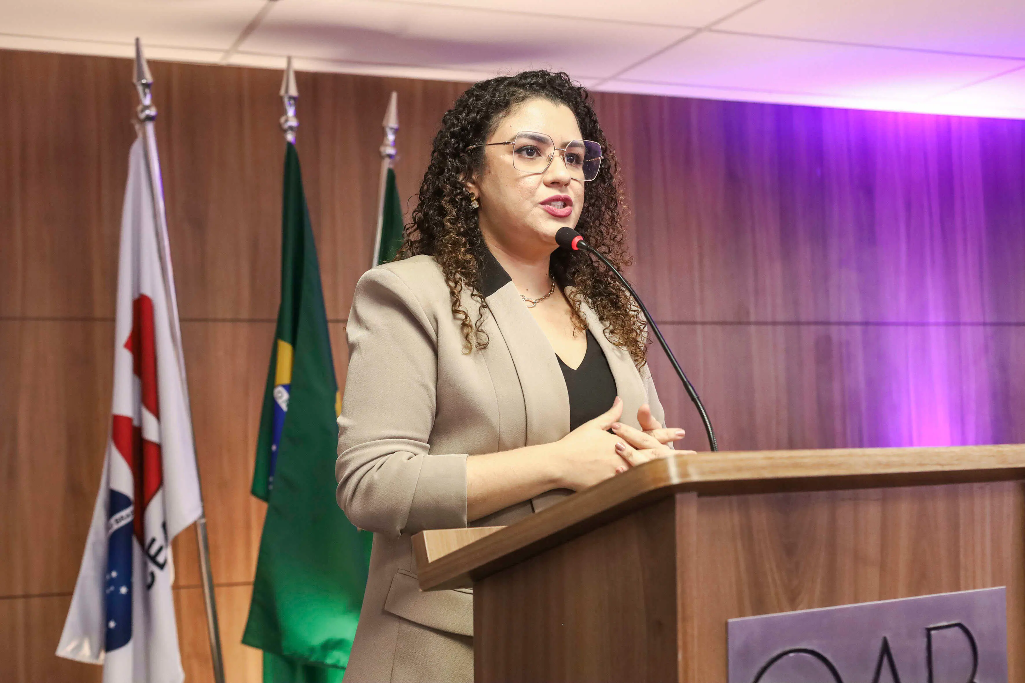 Mulher de cabelos cacheados e óculos fala ao microfone em um púlpito de madeira, em um ambiente institucional. Ela veste blazer bege e blusa preta, com as mãos juntas à frente do corpo. Ao fundo, há bandeiras — incluindo a do Brasil — e uma parede de madeira, com iluminação suave em tons de roxo.