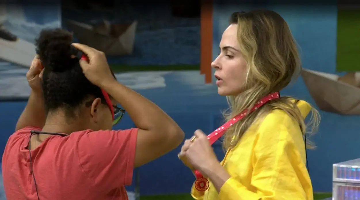 Duas pessoas em um ambiente interno; uma ajusta o cabelo enquanto a outra segura uma fita vermelha, com cenário pintado ao fundo.