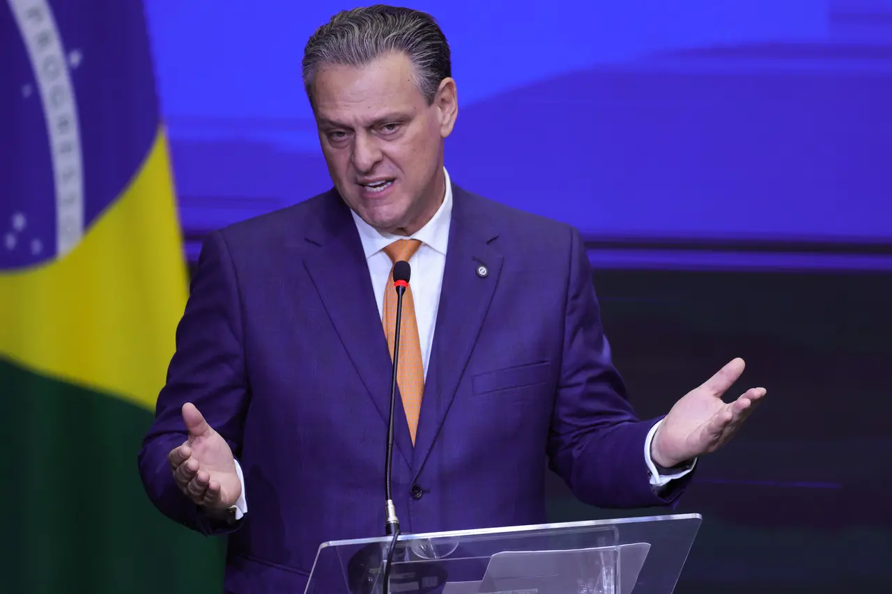 Pessoa em pé atrás de um púlpito transparente, usando terno escuro e gravata, com microfone à frente e mãos abertas em gesto de explicação, diante de fundo azul e bandeira ao lado.