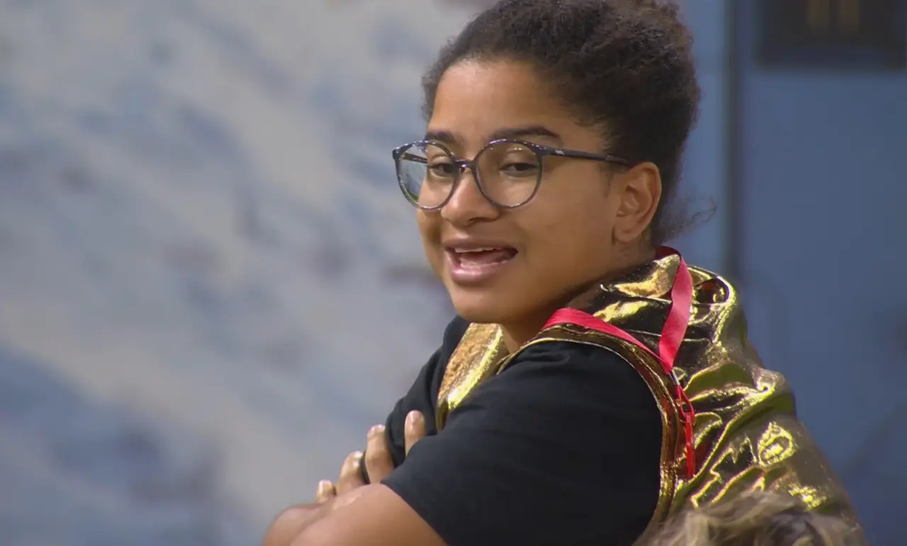 Milena, mulher negra de cabelo crespo amarrado em coque e usando óculos de grau redondo, blusa preta, colete dourado e cordão vermelho.