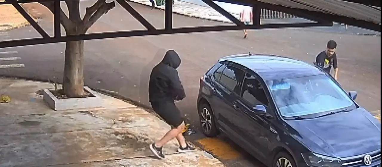 Imagem de câmera de segurança aérea mostrando carro preto, onde entram uma pessoa de capuz e um rapaz de camisa preta e cabelo preto.