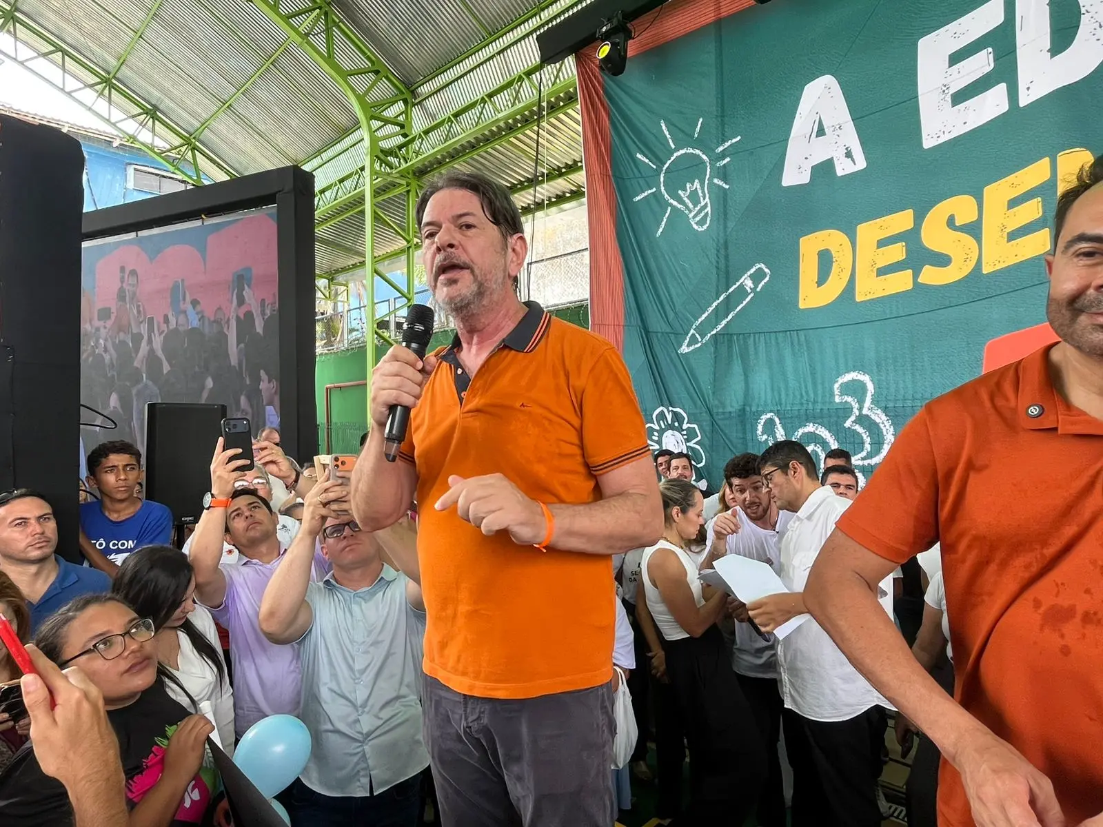 Homem branco de barba e cabelos claros, meio brancos, usando camisa polo laranja.