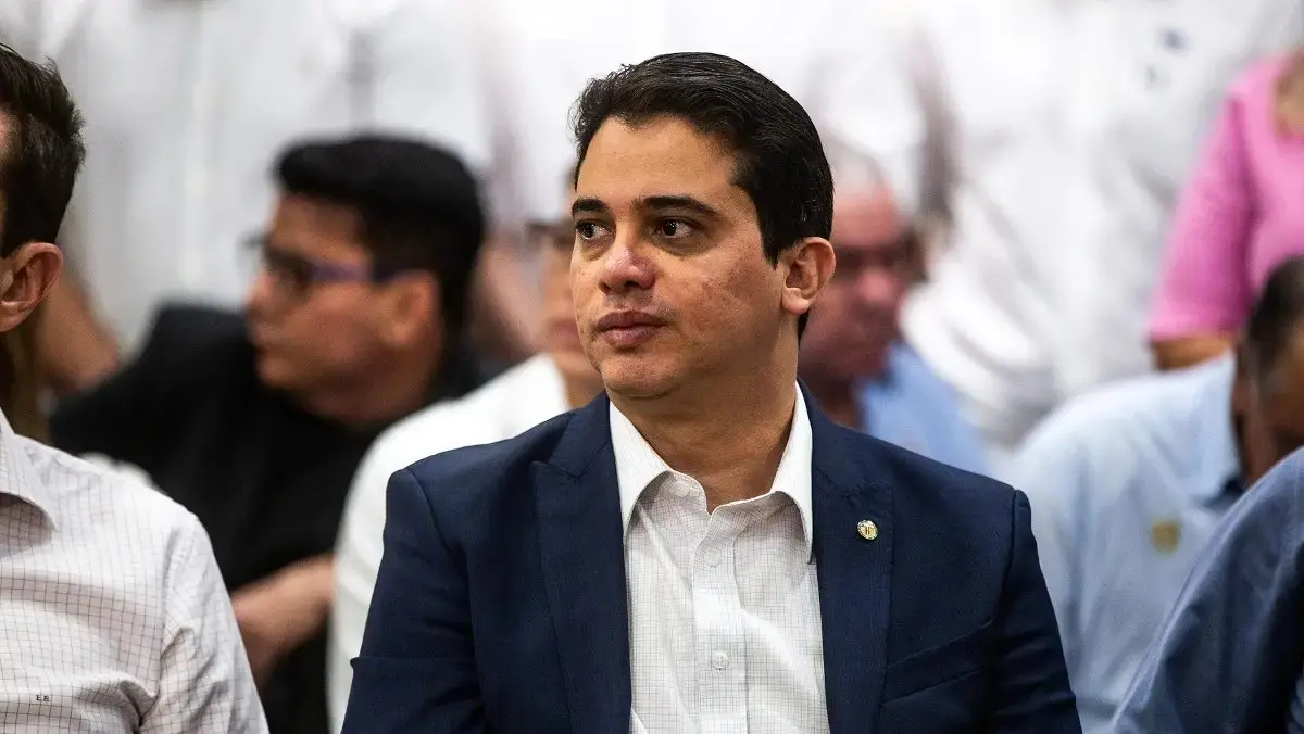 Homem de cabelos escuros, usando terno azul e camisa branca, sentado em um ambiente interno com outras pessoas ao fundo desfocadas, olhando para o lado com expressão séria.