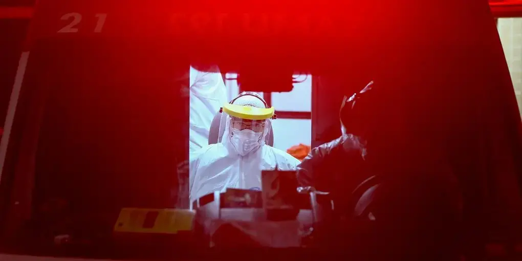 Profissional da saúde é fotografado no interior de uma ambulância. O profissional está protegido com roupas especiais e um face shield.