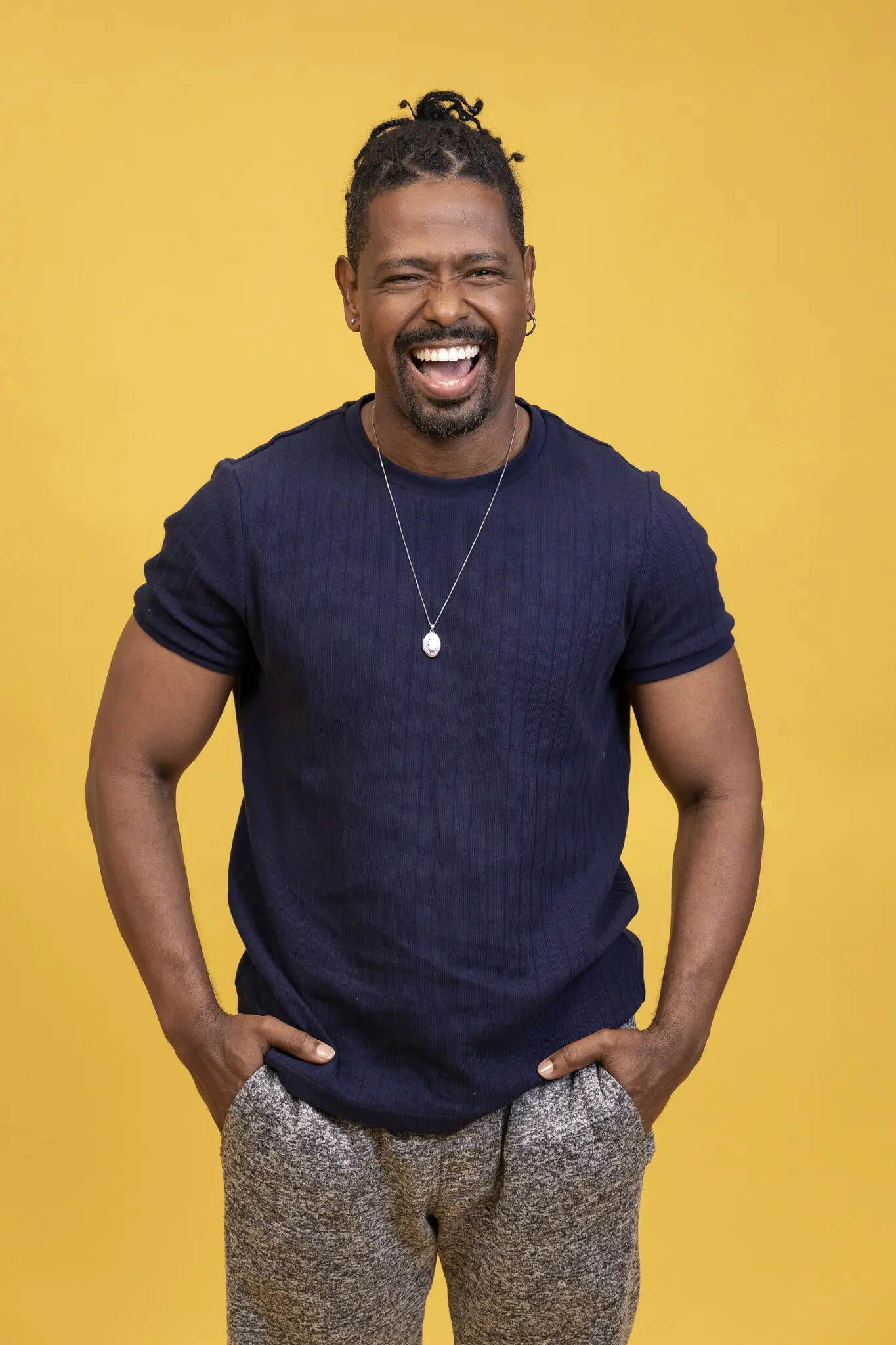 Retrato de Leandro, sorrindo, com o cabelo amarrado, usando blusa azul marinho e em frente a fundo amarelo.