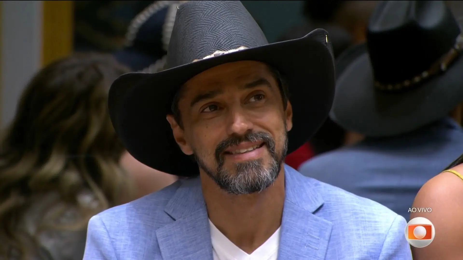 Homem de meia-idade, com cabelos escuros e grisalhos e barba por fazer, sorri enquanto olha para alguém fora do enquadramento. Ele veste uma camiseta preta e um cordão com credencial no pescoço. Ao fundo, há outras pessoas e elementos de um ambiente interno, possivelmente um estúdio ou casa com iluminação artificial.