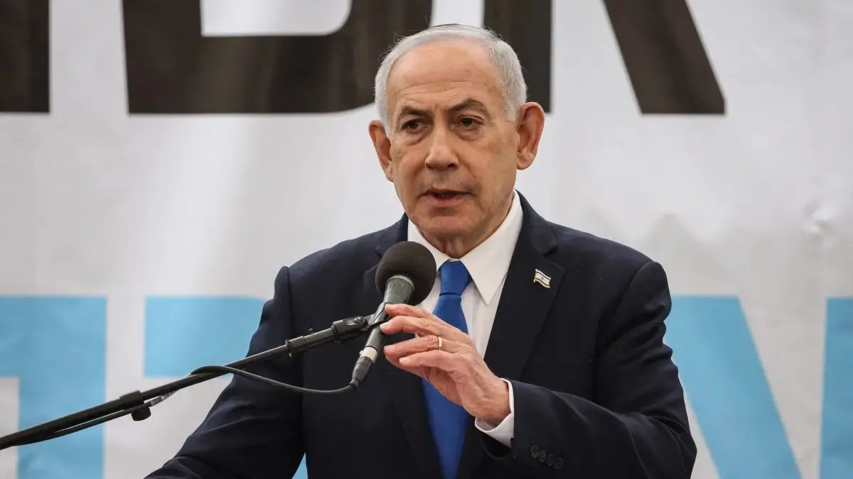 Homem de terno escuro e gravata azul fala ao microfone em um púlpito, com a bandeira de Israel presa na lapela. Ao fundo, há um painel claro com letras grandes parcialmente visíveis. Ele gesticula com a mão enquanto discursa, em um ambiente que parece ser um evento oficial ou pronunciamento público.