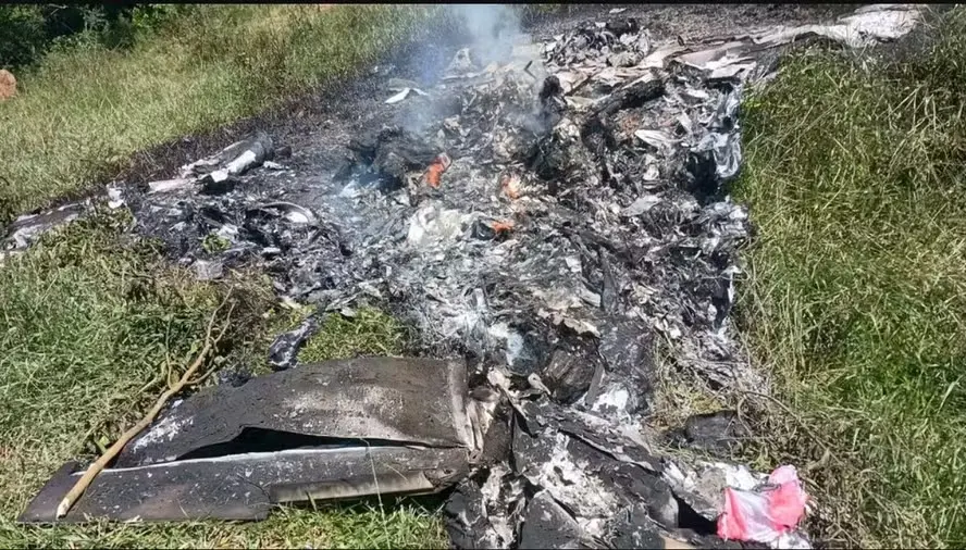 Destroços de uma aeronave espalhados em uma área de vegetação, com partes completamente queimadas e ainda soltando fumaça. Entre os restos carbonizados, é possível ver fragmentos metálicos retorcidos e objetos parcialmente destruídos sobre a grama. O local aparenta ser uma área rural, com vegetação ao redor do ponto de queda.