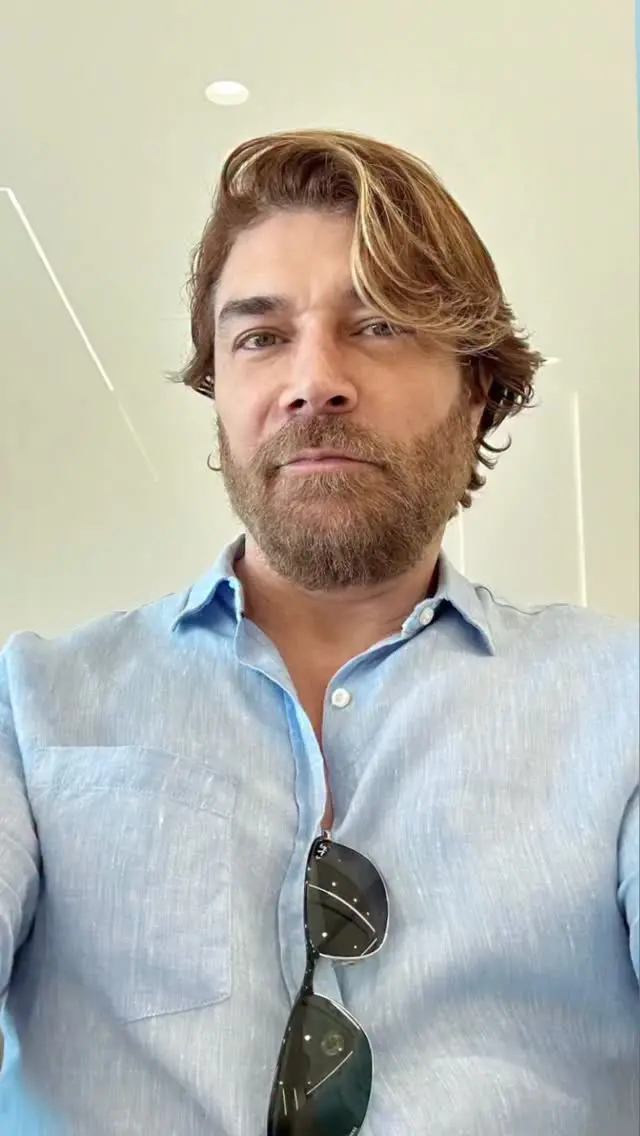 Homem de cabelo castanho claro, barba bem cuidada, olhando sério para a câmera, usando camisa azul claro e óculos escuros pendurados na camisa, em ambiente indoor bem iluminado.