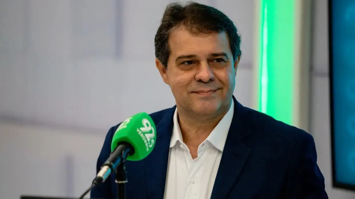 Homem sorridente falando ao microfone em entrevista de rádio ou podcast, cenário moderno com cores azul e verde ao fundo, profissional e confiante.