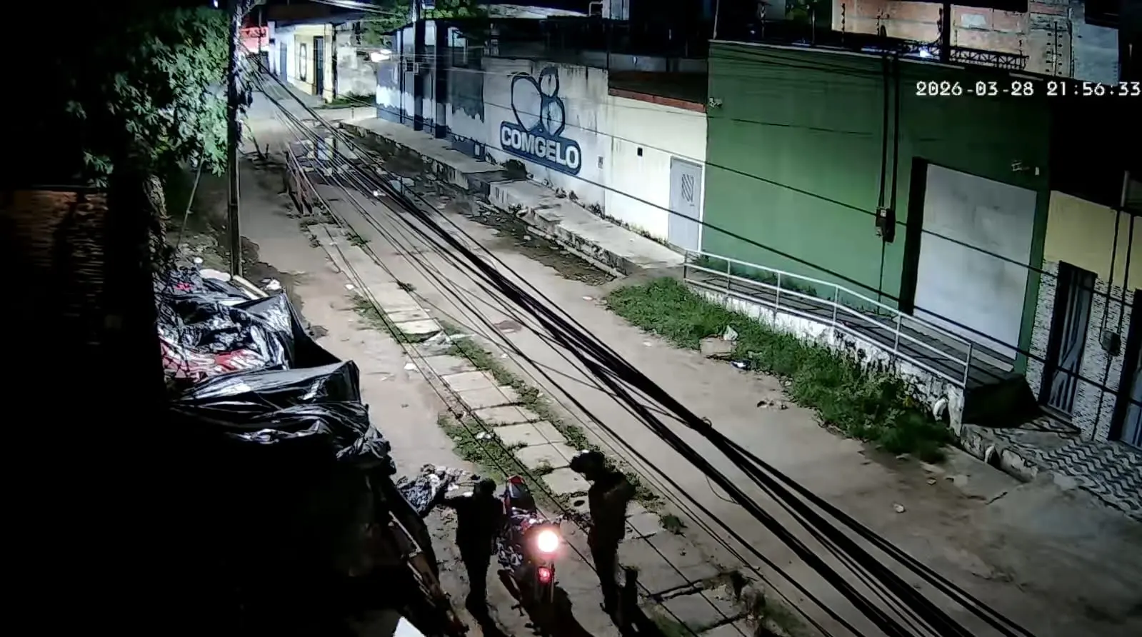 Imagem de uma rua urbana à noite com cabos de energia visíveis, um grupo de policiais e um motociclista com farol aceso, ao lado de uma construção com publicidade de congelados, sem pessoas reconhecíveis ou elementos específicos que indiquem uma celebridade ou personagem famoso.