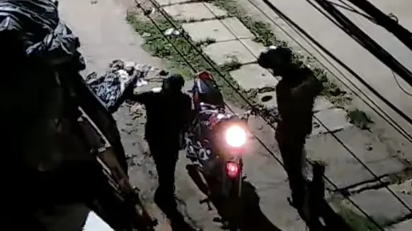 Imagem de uma rua urbana à noite com cabos de energia visíveis, um grupo de policiais e um motociclista com farol aceso, ao lado de uma construção com publicidade de congelados, sem pessoas reconhecíveis ou elementos específicos que indiquem uma celebridade ou personagem famoso.