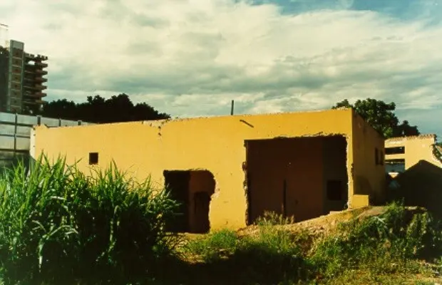 Construção baixa de paredes amarelas parcialmente destruídas, com aberturas sem portas, vegetação alta ao redor e prédios ao fundo.