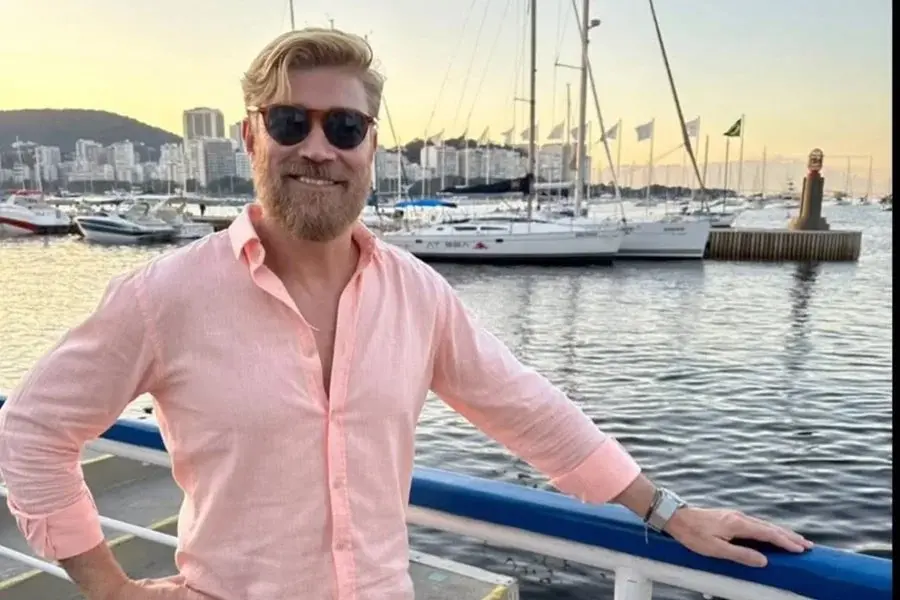 Homem com barba e óculos escuros sorrindo na orla do porto com barcos ao fundo ao entardecer, roupas casuais, clima de lazer e relaxamento.