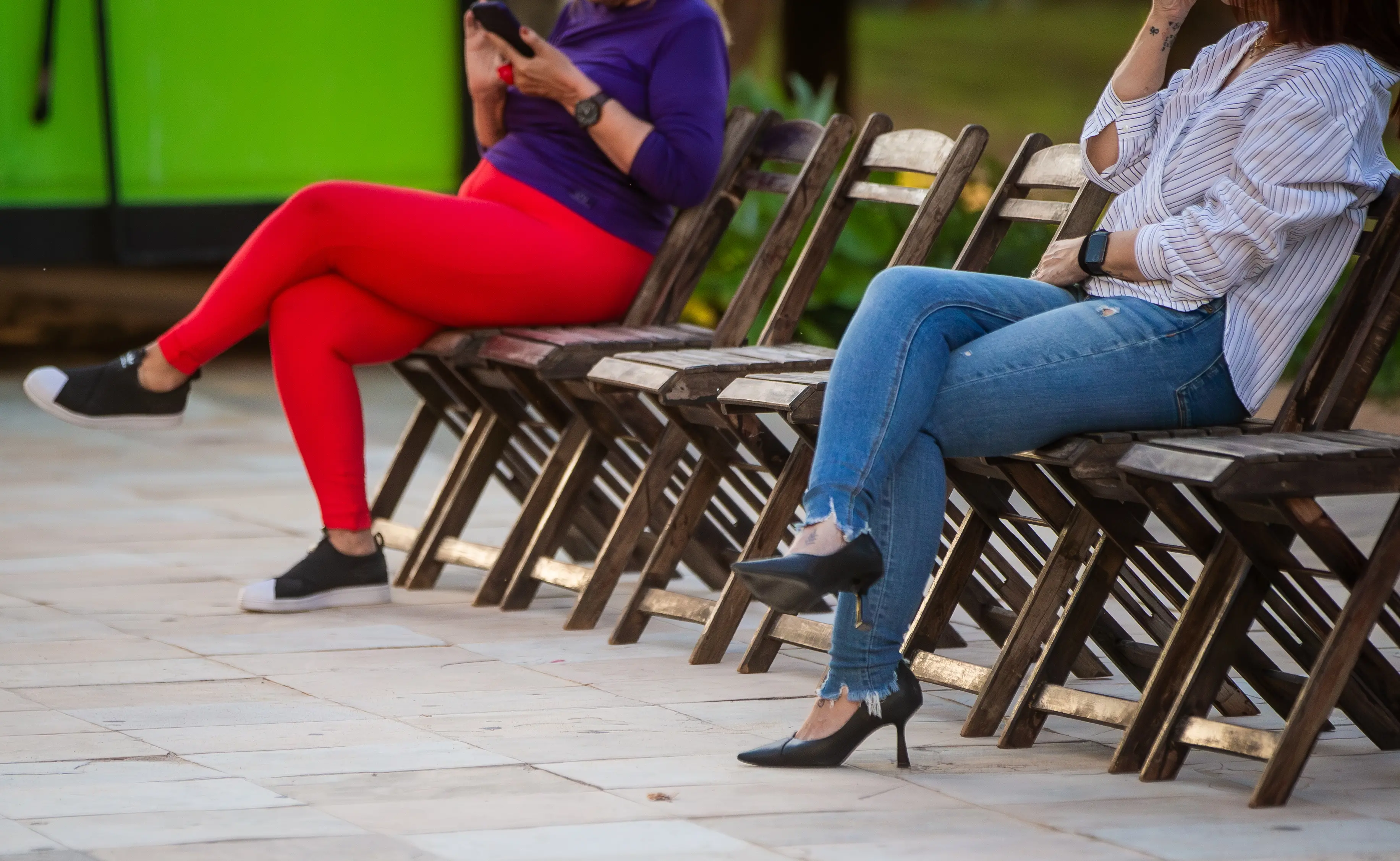 Duas mulheres sentadas em cadeiras enfileradas. O foco da imagem é da cintura para baixo. As pernas estão em evidência.