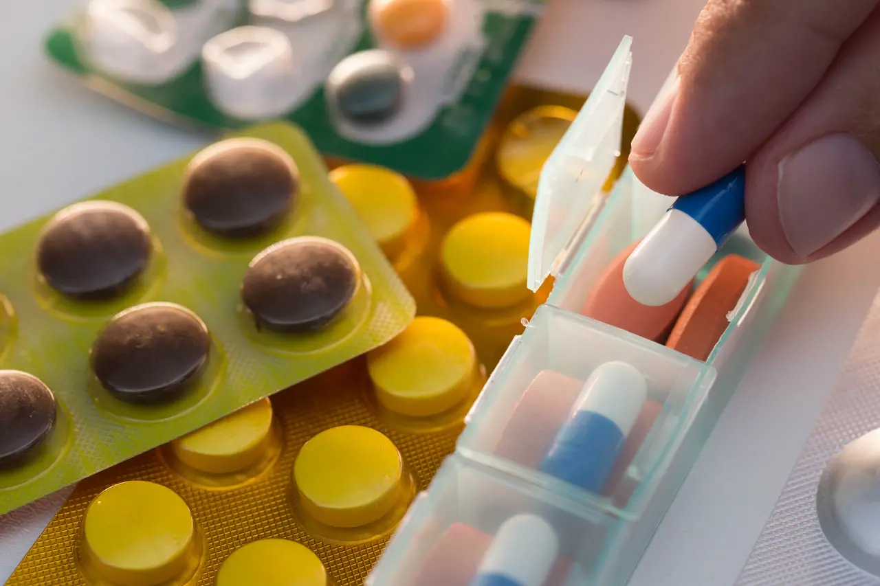 Uma mão segura uma cápsula azul e branca próxima a um organizador semanal de medicamentos, cercado por várias cartelas de comprimidos coloridos em tons de amarelo e marrom. A cena destaca a organização da rotina de saúde com foco em cores vibrantes e iluminação suave sobre os blisters.