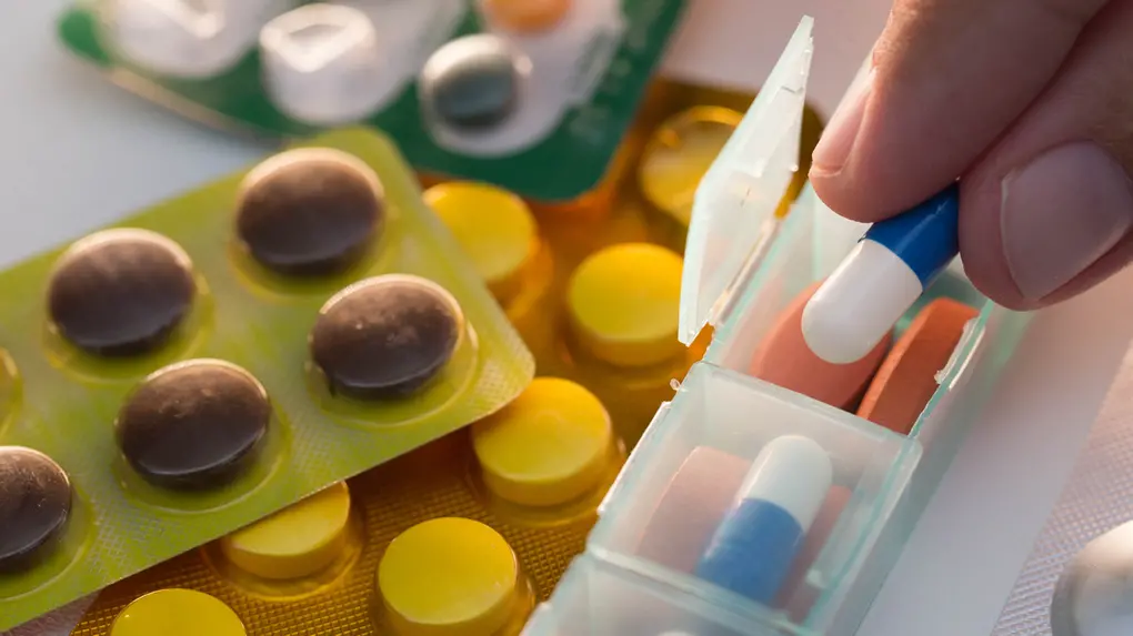 Uma mão segura uma cápsula azul e branca próxima a um organizador semanal de medicamentos, cercado por várias cartelas de comprimidos coloridos em tons de amarelo e marrom. A cena destaca a organização da rotina de saúde com foco em cores vibrantes e iluminação suave sobre os blisters.