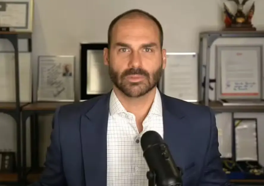 Homem de negócios sério com barba e cabelo raspado, vestindo terno azul claro, falando em frente à câmera com várias certificações e prêmios ao fundo.