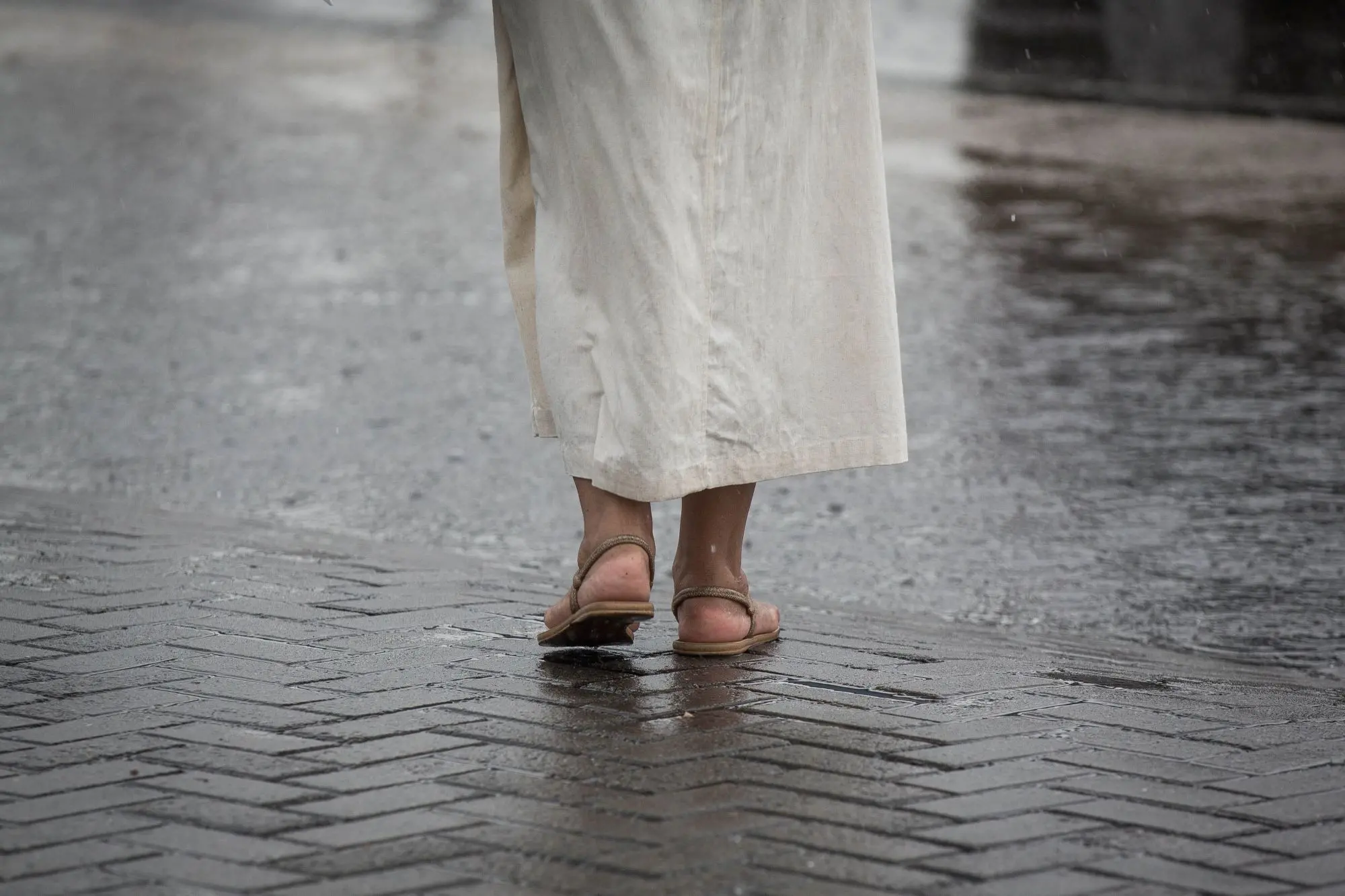 Pessoa vista de trás caminhando em uma rua molhada, usando sandálias e uma saia longa clara, com o chão refletindo a umidade da chuva.