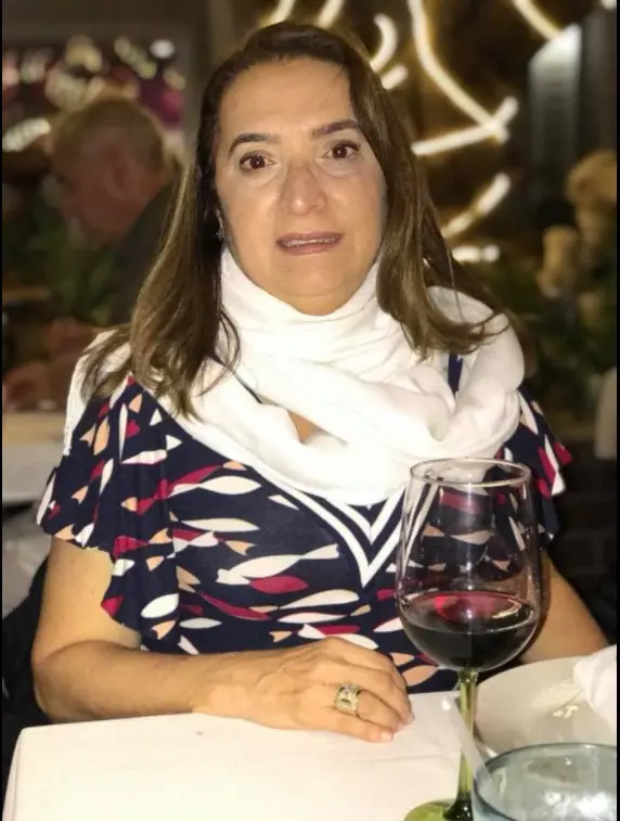 Mulher sentada à mesa de um restaurante, olhando para a câmera. Ela tem cabelos castanhos na altura dos ombros e usa um vestido estampado em azul, branco e vermelho, com um lenço branco no pescoço. À sua frente, sobre a mesa, há uma taça de vinho tinto. O ambiente ao fundo tem iluminação quente e outros clientes desfocados. A mulher é a médica Lúcia Belém, que morreu atropelada em Fortaleza em 2021.