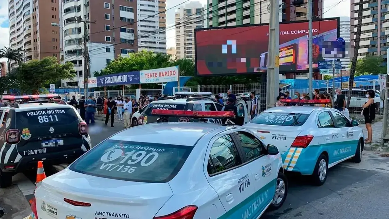 Rua em Fortaleza com vários carros da Polícia Militar e da AMC Trânsito estacionados, alguns com luzes ligadas. Há um grupo de pessoas reunidas atrás de grades, próximo a uma estrutura de testagem rápida de Covid‑19. Ao fundo, aparecem prédios altos e um grande painel eletrônico. No local ocorreu o atropelamento da médica Lúcia Belém.