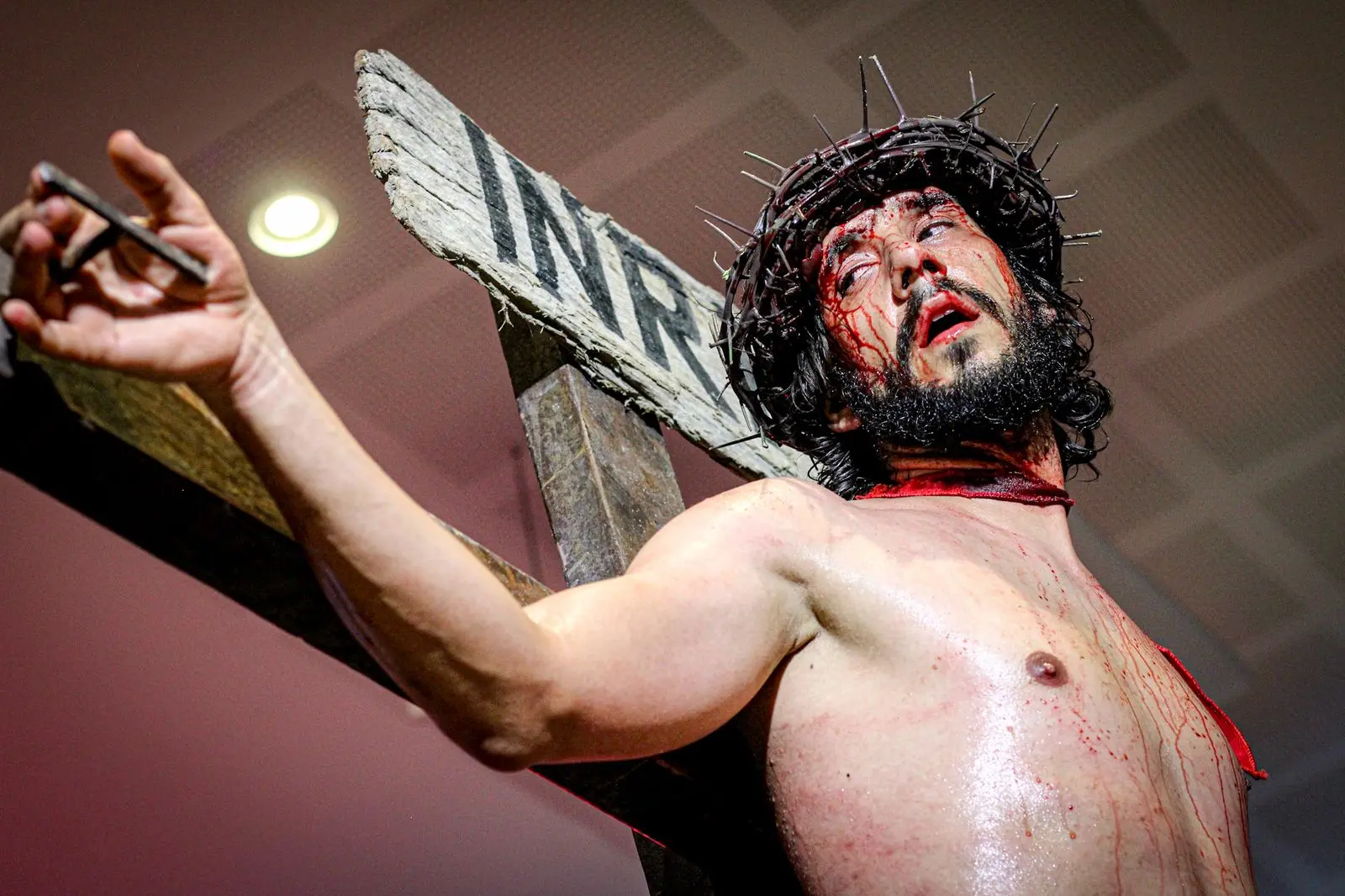 Um ator interpreta Jesus Cristo crucificado no espetáculo 'Paixão de Cristo', usando uma coroa de espinhos e maquiagem que simula sangue no rosto e peito. A cena, capturada em um ângulo baixo, destaca a placa 'INRI' na cruz de madeira sob uma iluminação dramática que enfatiza a carga emocional da encenação.