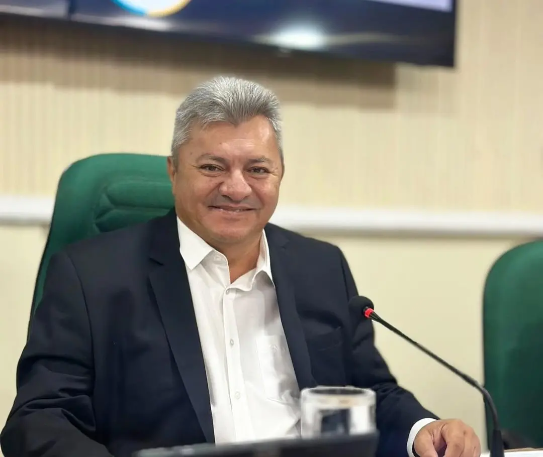 Homem de meia-idade, com cabelos grisalhos e expressão sorridente, sentado em uma cadeira verde em ambiente formal, possivelmente uma sala de reuniões ou plenário. Ele veste terno escuro com camisa branca e está posicionado diante de um microfone de mesa com luz vermelha acesa. À sua frente há um copo com água. Ao fundo, há uma parede clara e parte de uma tela ou monitor desfocado.