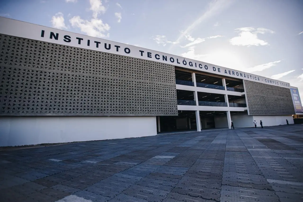 Fachada moderna do Instituto Tecnológico de Aeronáutica (Campus Ceará), com painéis vazados geométricos, estrutura de vários andares aberta ao centro, amplo pátio pavimentado à frente e algumas pessoas caminhando sob céu parcialmente nublado.