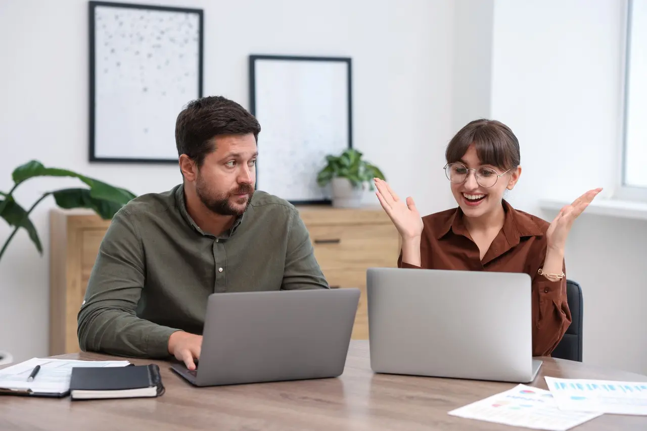 Homem e mulher trabalham em laptops; ela comemora com as mãos erguidas enquanto ele observa ao lado, em escritório.