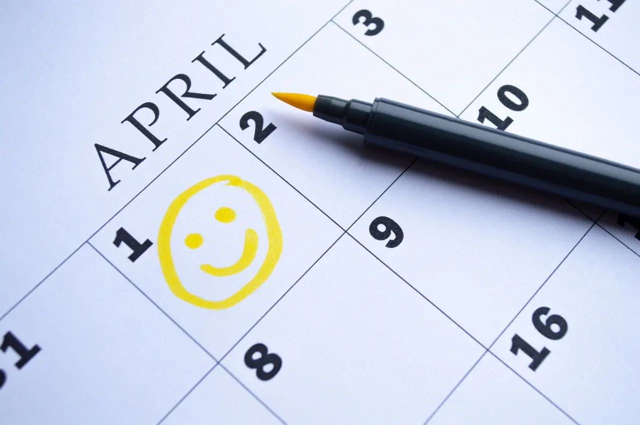 Um calendário de papel exibe o mês de abril com uma carinha sorridente desenhada em amarelo sobre o dia 1. Uma caneta hidrográfica de ponta amarela repousa sobre a folha, destacando a marcação vibrante que celebra o Dia da Mentira.