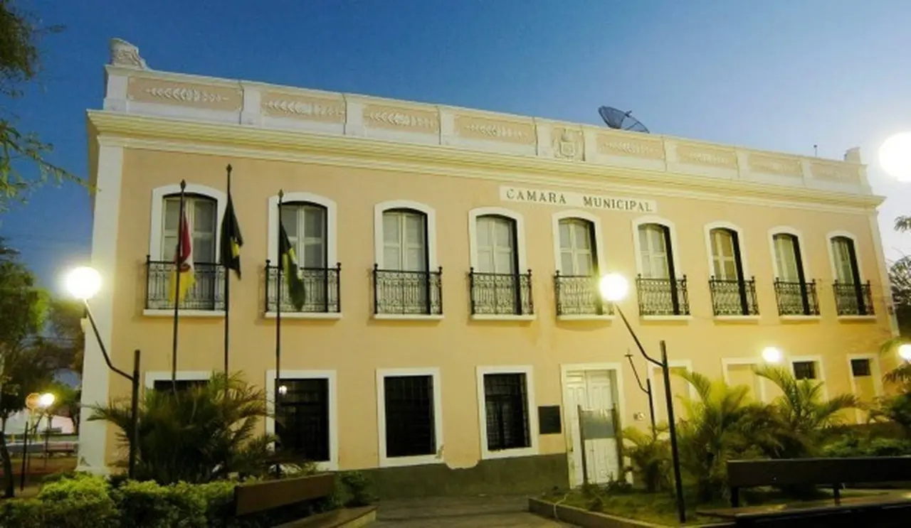 Fachada de prédio na cor amarela, com nove pequenas sacadas no andar de cima e oito janelas no andar inferior, além de uma porta aberta ao centro, durante o período noturno.