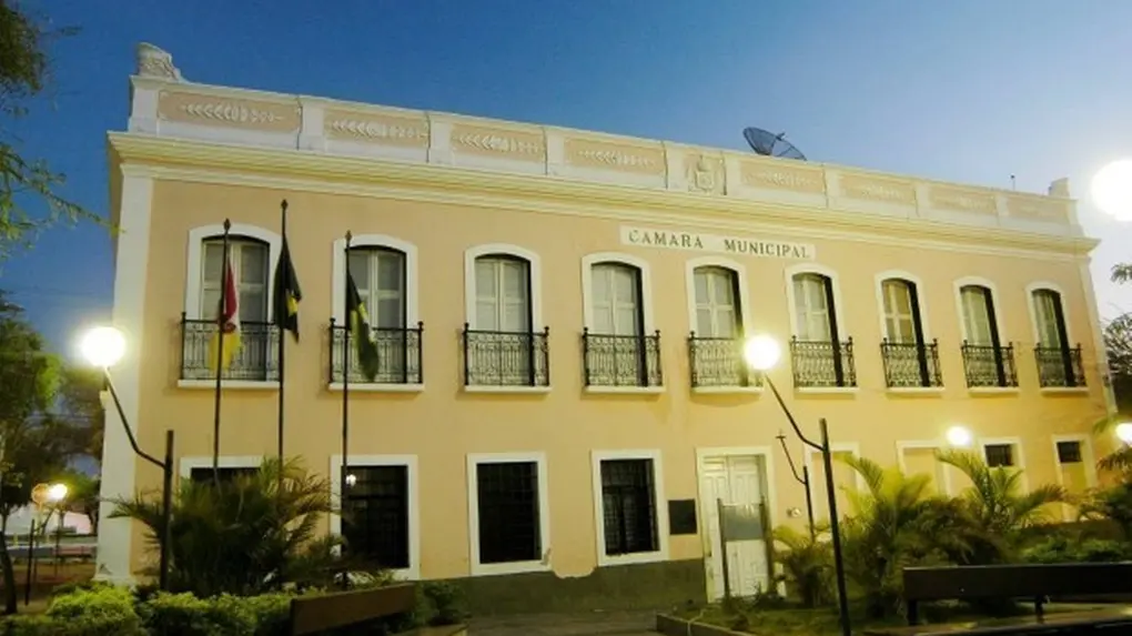 Fachada de prédio na cor amarela, com nove pequenas sacadas no andar de cima e oito janelas no andar inferior, além de uma porta aberta ao centro, durante o período noturno.