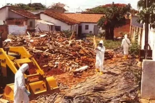 Técnicos com roupas de proteção brancas e máscaras trabalham em um terreno coberto por lonas escuras, cercado por escombros e uma retroescavadeira amarela. Ao fundo, casas simples e árvores compõem o cenário da operação de limpeza e descontaminação em uma área residencial.