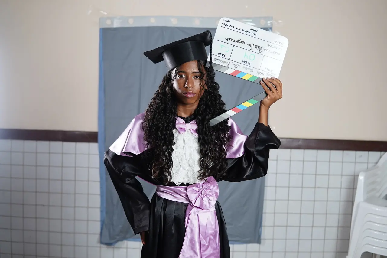 Uma jovem de cabelos longos e cacheados posa vestindo uma beca de formatura preta com detalhes em cetim lilás e capelo. Ela segura uma claquete de cinema aberta na altura do rosto, posicionada contra um fundo simples composto por uma cortina cinza e parede de azulejos brancos.