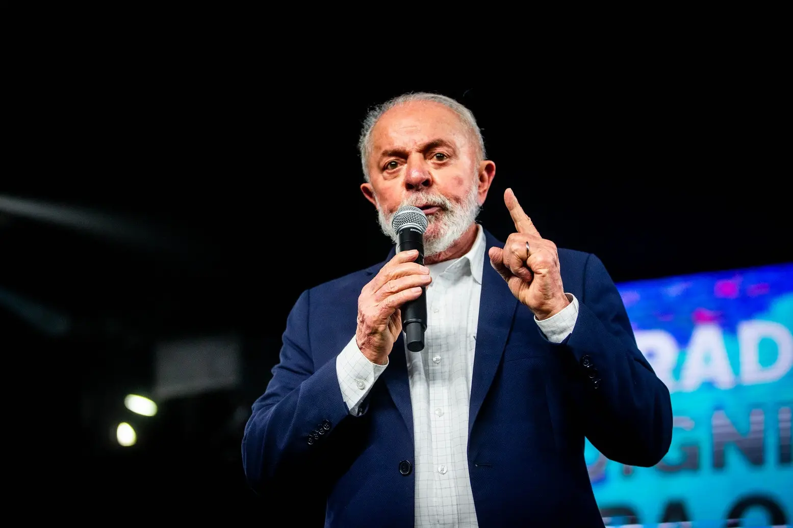 Presidente Lula em terno azul discursa em palanque segurando microfone e com dedo indicador erguido.