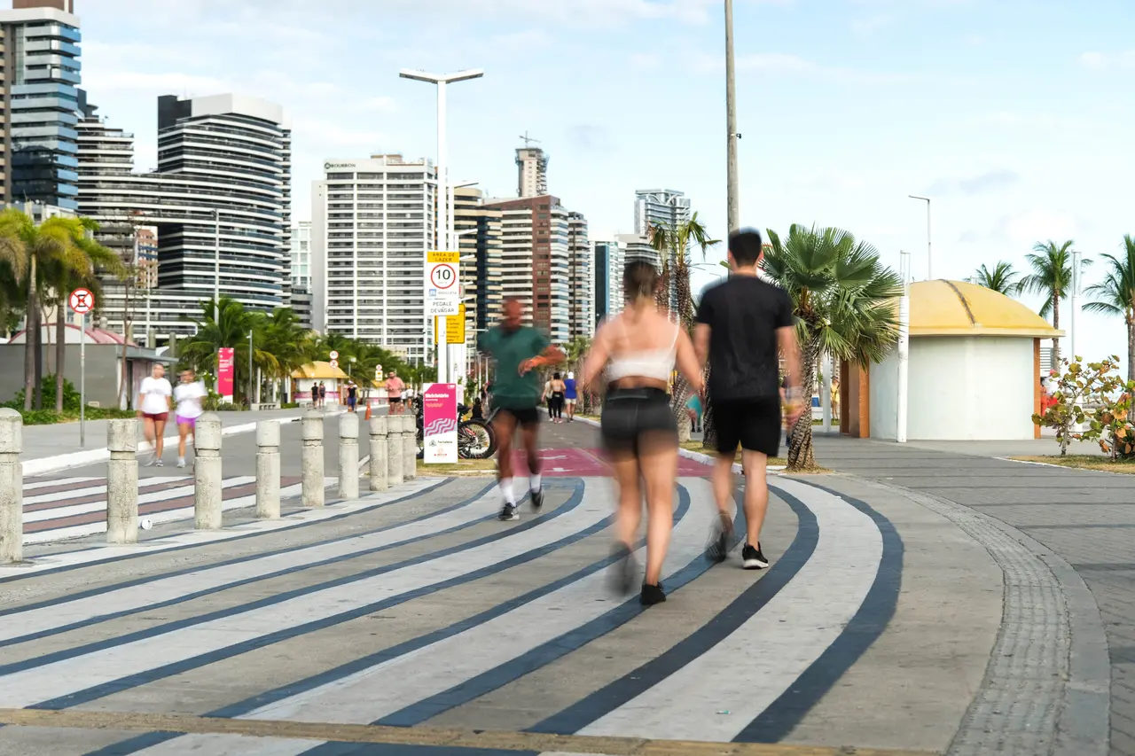 Pessoas caminhando e correndo na orla de Fortaleza, em uma via compartilhada com faixas curvas, prédios altos ao fundo, palmeiras e quiosques à beira-mar em dia claro.