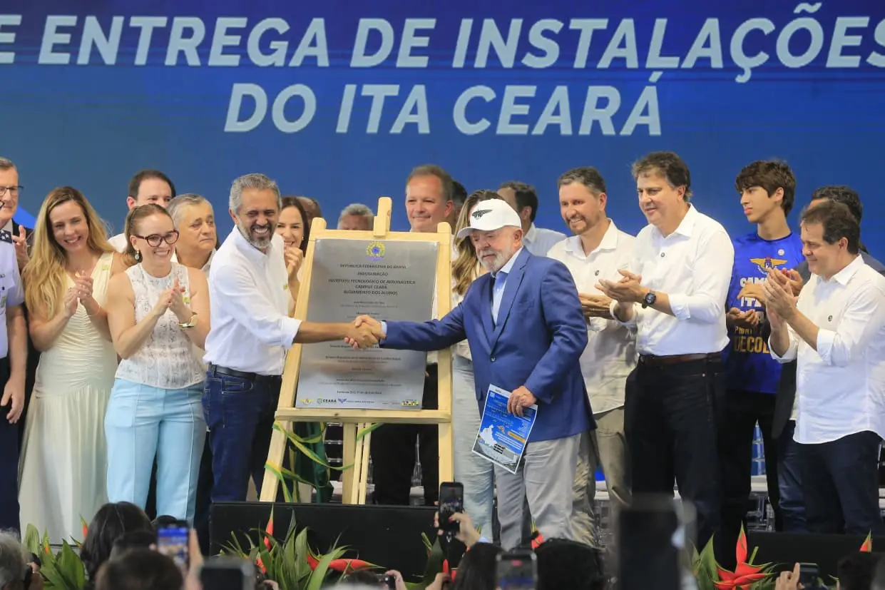 Lula e o governador Elmano de Freitas apertam as mãos ao lado de uma placa de inauguração do ITA Ceará. Eles estão cercados por diversas autoridades que aplaudem o ato em um palco decorado.