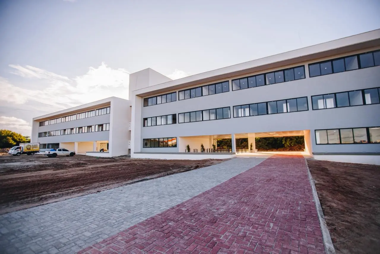Fachada externa de um edifício moderno de três andares com janelas horizontais e acabamento cinza e branco. À frente, há um caminho de bloquetes intertravados em tons de cinza e vermelho.