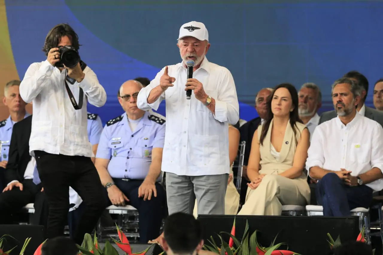 O presidente Lula, de camisa branca e boné, discursa ao microfone em um palco no ITA Ceará. Ele aponta para frente enquanto é observado por autoridades sentadas ao fundo e um fotógrafo em primeiro plano.