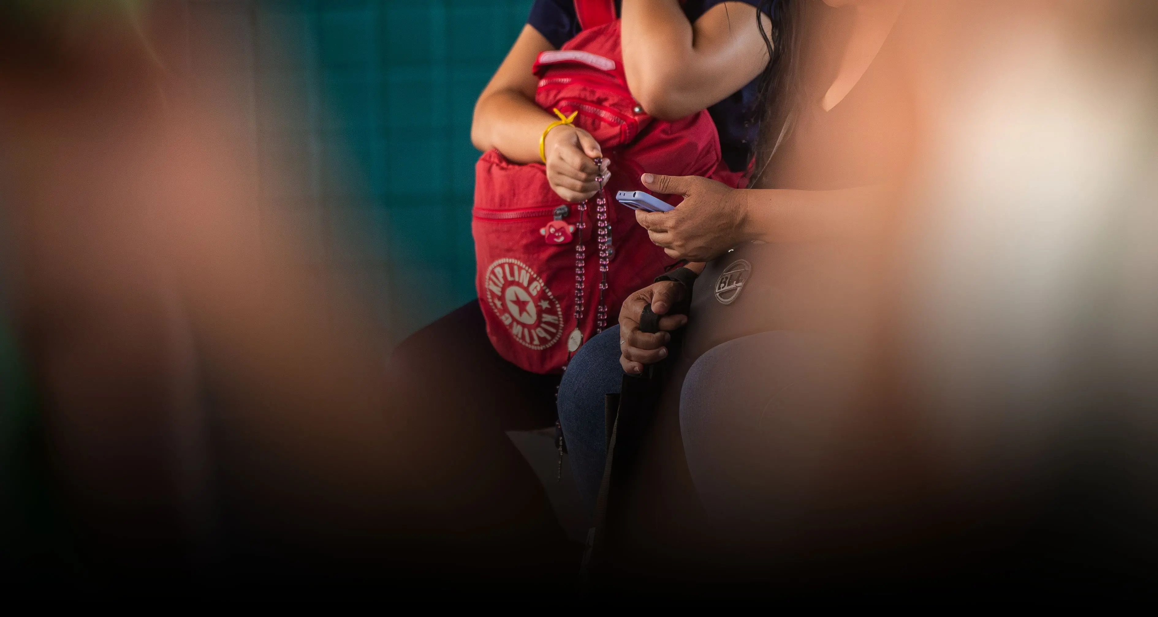 Pessoas sentadas em local público; uma delas segura um celular azul e a outra segura um terço sobre uma mochila vermelha. A cena tem foco seletivo com elementos desfocados em primeiro plano.