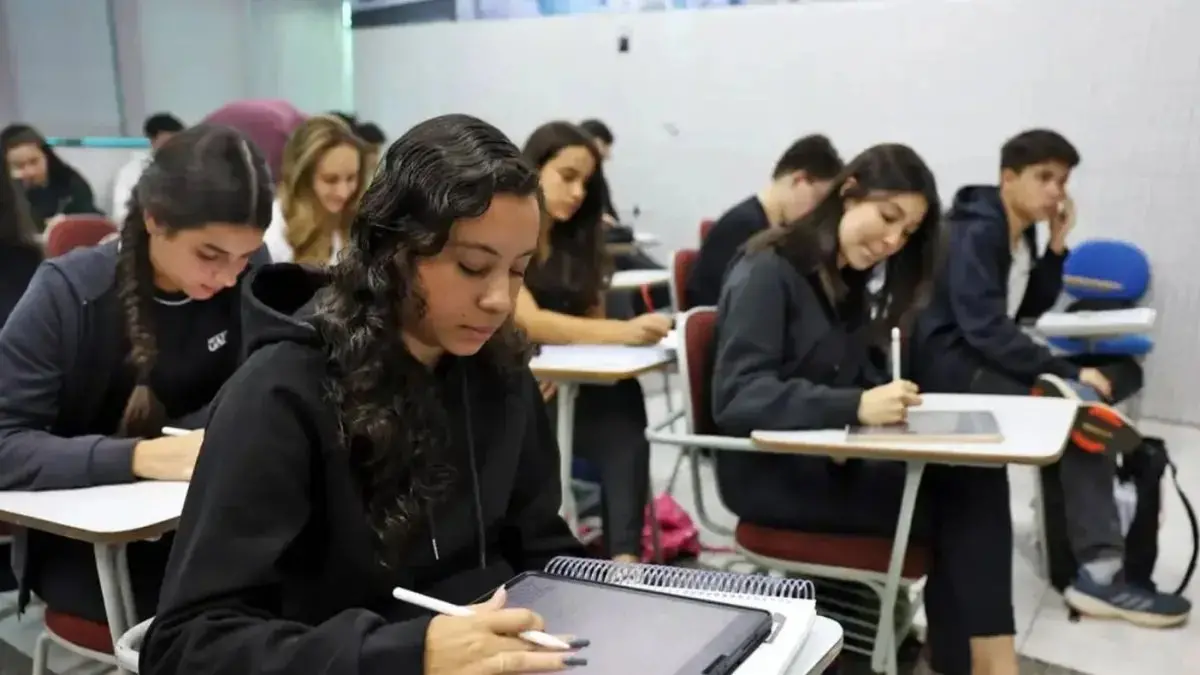 Imagem mostra grupo de estudantes em sala de aula de escola brasileira, usando roupas escuras, focados em suas atividades, com um ambiente organizado e educativo.