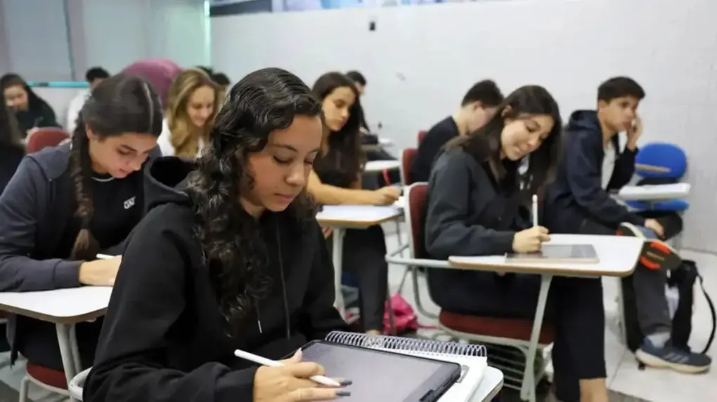 Imagem mostra grupo de estudantes em sala de aula de escola brasileira, usando roupas escuras, focados em suas atividades, com um ambiente organizado e educativo.